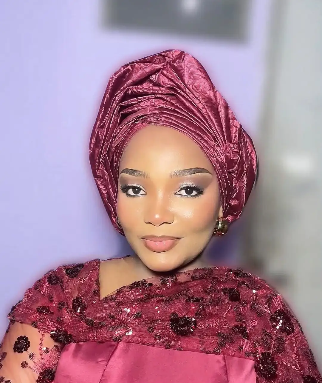 A smiling guest in a wine gele and sequinned lace, with soft pink glam